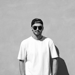 Ryan Gregory, a man wearing a light t-shirt, a cap on backwards, and sunglasses, depicted in a black and white headshot with a blank background that is actually a yellow coloured wall in Venice, Italy